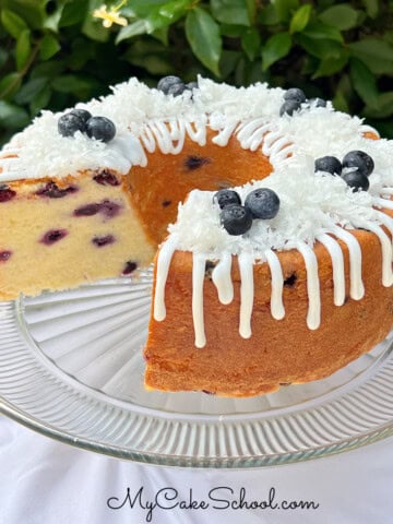 Blueberry Coconut Pound Cake, sliced, on a pedestal. It is topped with a coconut glaze, flaked coconut, and fresh blueberries.